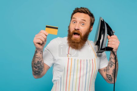 impressed bearded man with open mouth holding iron and credit card isolated on blue