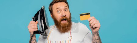 Foto de Hombre barbudo asombrado mostrando hierro y tarjeta de crédito aislado en azul, bandera - Imagen libre de derechos