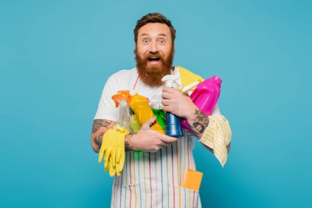 amazed and happy man holding various detergents and rubber gloves while looking at camera isolated on blue