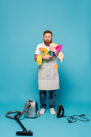 full length of serious tattooed man holding detergents near vacuum cleaner and iron on blue background