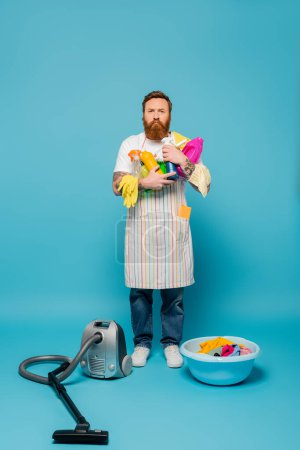 full length of serious man holding detergents near vacuum cleaner and iron on blue background