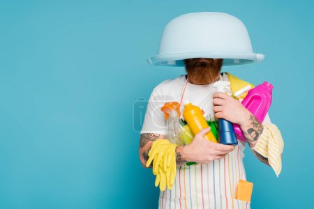 bearded man with different cleaning supplies and laundry bowl on head isolated on blue