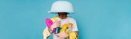 Photo for Tattooed man with cleaning supplies and laundry bowl on head isolated on blue, banner - Royalty Free Image