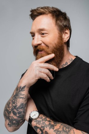 happy bearded man in black t-shirt and wristwatch holding tattooed hand near face isolated on grey