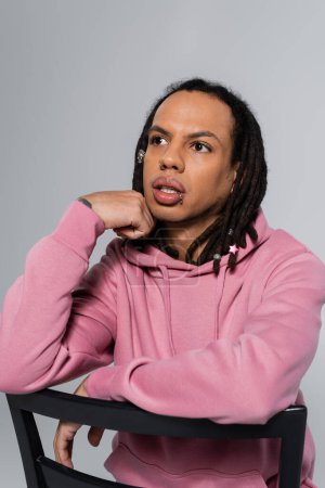 thoughtful multiracial man in pink hoodie leaning on chair back isolated on grey 