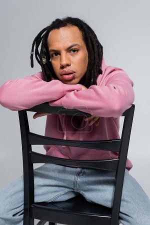 pierced multiracial man in pink hoodie sitting on chair and looking at camera isolated on grey 