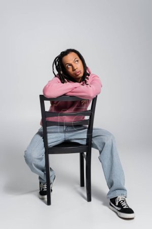 full length of pensive multiracial man with dreadlocks sitting on chair on grey 