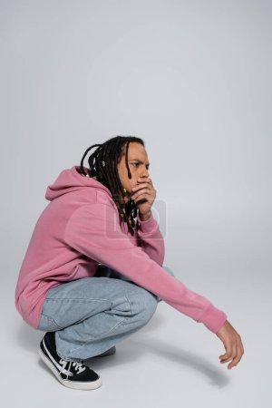 Photo for Full length of worried multiracial man with dreadlocks sitting on haunches and covering mouth on grey - Royalty Free Image