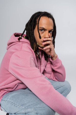 worried multiracial man with dreadlocks sitting on haunches and covering mouth isolated on grey 