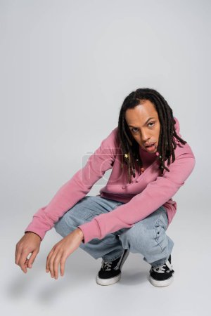 full length of multiracial man with dreadlocks sitting on haunches and looking at camera on grey 