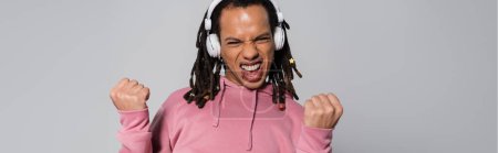 excited multiracial man with dreadlocks listening music in wireless headphones and showing yes gesture isolated on grey, banner 
