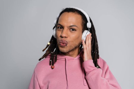 Téléchargez les photos : Multiracial man with dreadlocks pouting lips while listening music in wireless headphones isolated on grey - en image libre de droit