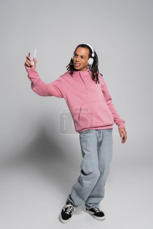 full length of multiracial man with dreadlocks taking selfie while listening music in wireless headphones on grey 