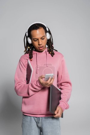 multiracial man in wireless headphones using smartphone and holding devices on grey 