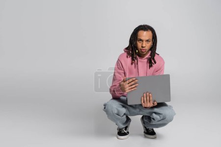 multiracial man in pink hoodie holding laptop while sitting on haunches on grey 