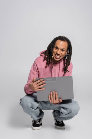 happy multiracial man in pink hoodie holding laptop while sitting on haunches on grey 