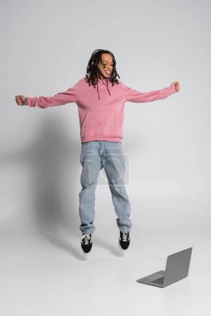 Photo for Full length of happy multiracial man in pink hoodie and jeans jumping near laptop on grey - Royalty Free Image