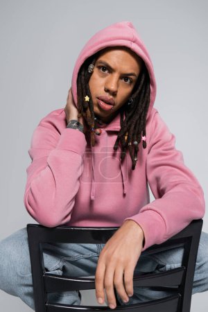portrait of tattooed multiracial man in pink hoodie looking at camera while leaning on chair back isolated on grey 