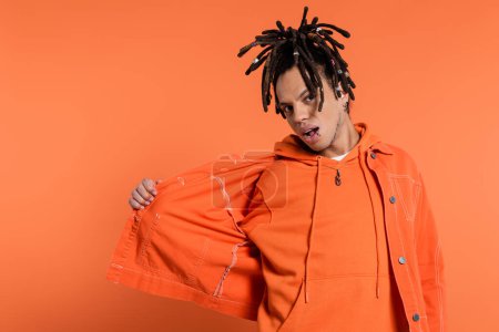multiracial man with dreadlocks and opened mouth adjusting denim jacket on coral background 