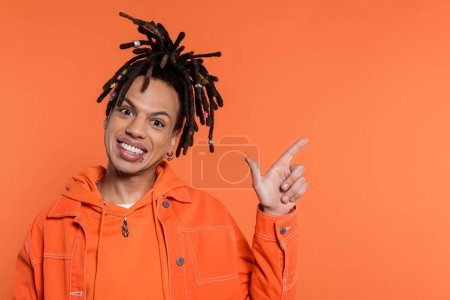 happy multiracial man with dreadlocks pointing with finger isolated on coral background 
