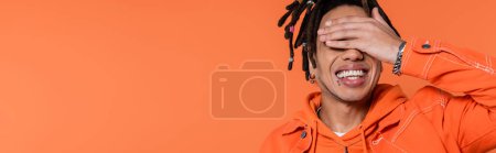 happy multiracial man with dreadlocks and tattoo covering eyes isolated on coral background, banner 