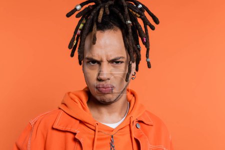 Photo for Sad multiracial man with dreadlocks pouting lips isolated on coral background - Royalty Free Image