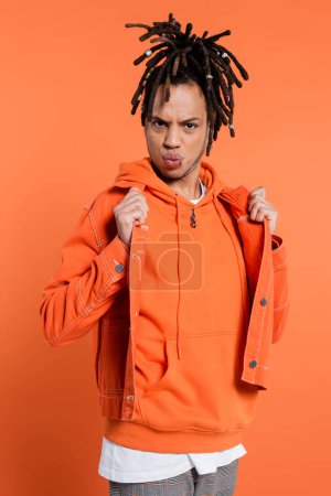 multiracial man with dreadlocks adjusting denim jacket while pouting lips on coral 