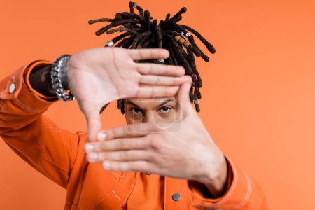 Foto de Multiracial man with dreadlocks showing frame gesture while looking at camera isolated on coral background - Imagen libre de derechos