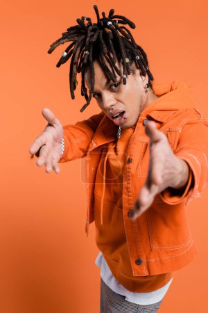 multiracial man with dreadlocks standing with outstretched hands while looking at camera on coral background 