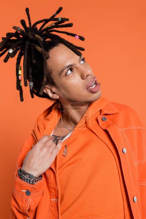 tattooed and multiracial man with dreadlocks adjusting hoodie while feeling hot isolated on coral 