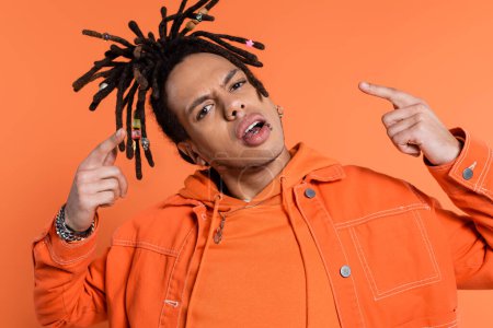 pierced and multiracial man with dreadlocks looking at camera while pointing with fingers isolated on coral background 