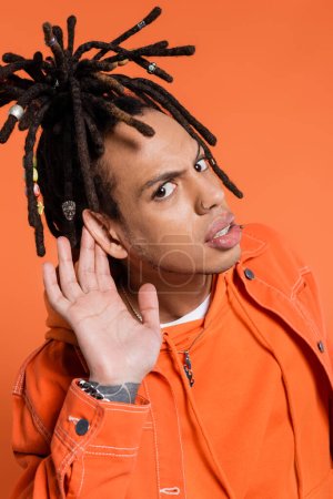 tattooed and multiracial man with dreadlocks looking at camera while touching ear isolated on coral 