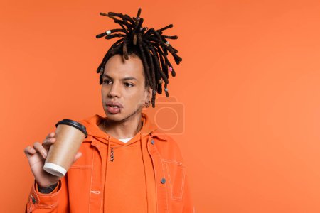 pensive and multiracial man with dreadlocks holding paper cup with coffee to go isolated on coral background 