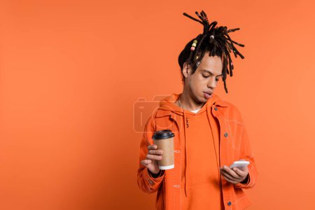 homme percé et multiracial avec dreadlocks tenant tasse en papier et en utilisant smartphone sur fond de corail 