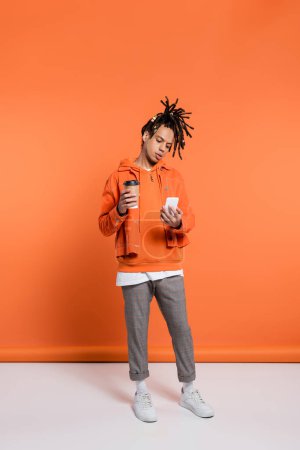 full length of pierced and multiracial man with dreadlocks holding paper cup and using smartphone on coral background 