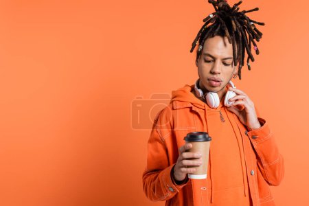 pierced and multiracial man with wireless headphones looking at paper cup with coffee to go on coral background 