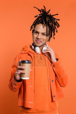 cheerful and multiracial man with wireless headphones holding paper cup with coffee to go isolated on coral 