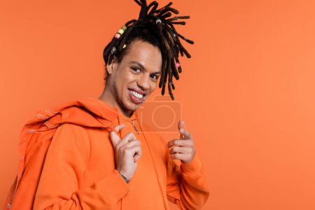 smiling multiracial man in hoodie holding denim jacket and pointing at camera isolated on coral background 
