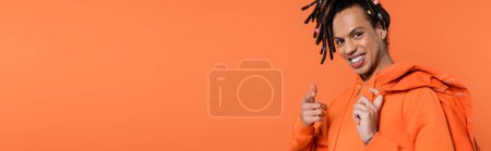 smiling multiracial man in hoodie holding denim jacket and pointing at camera isolated on coral background, banner 