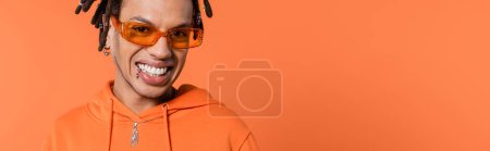 happy multiracial man with dreadlocks wearing stylish sunglasses isolated on coral background, banner 