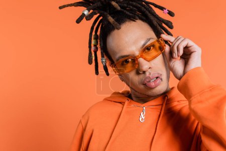 multiracial man with dreadlocks adjusting trendy sunglasses isolated on coral background 