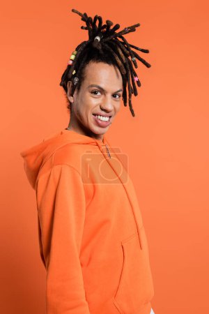 cheerful multiracial man with dreadlocks looking at camera and smiling isolated on coral 