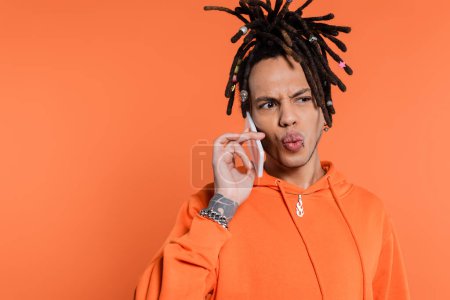 Photo for Tattooed multiracial man with dreadlocks pouting lips and talking on cellphone isolated on coral - Royalty Free Image