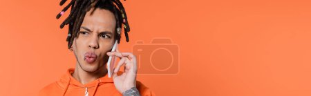 Photo for Multiracial man with dreadlocks pouting lips and talking on cellphone isolated on coral background, banner - Royalty Free Image