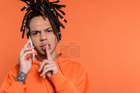 multiracial man with dreadlocks holding finger near lips and talking on cellphone isolated on coral 
