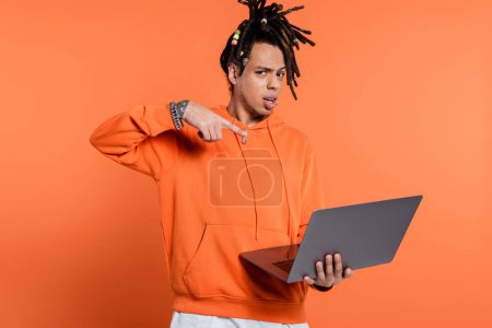 confused multiracial man with dreadlocks and tattoo pointing at laptop on coral background 