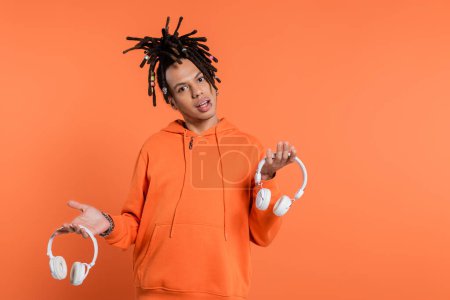 uncertain multiracial man with dreadlocks holding different wireless headphones and looking at camera on coral background 