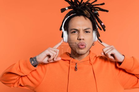 multiracial man with dreadlocks pointing at wireless headphones and pouting lips isolated on coral 
