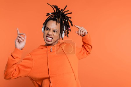 positive multiracial man listening music in wireless headphones and pointing with fingers on coral background