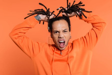 tattooed and emotional multiracial man standing with opened mouth and pulling dreadlocks on coral background